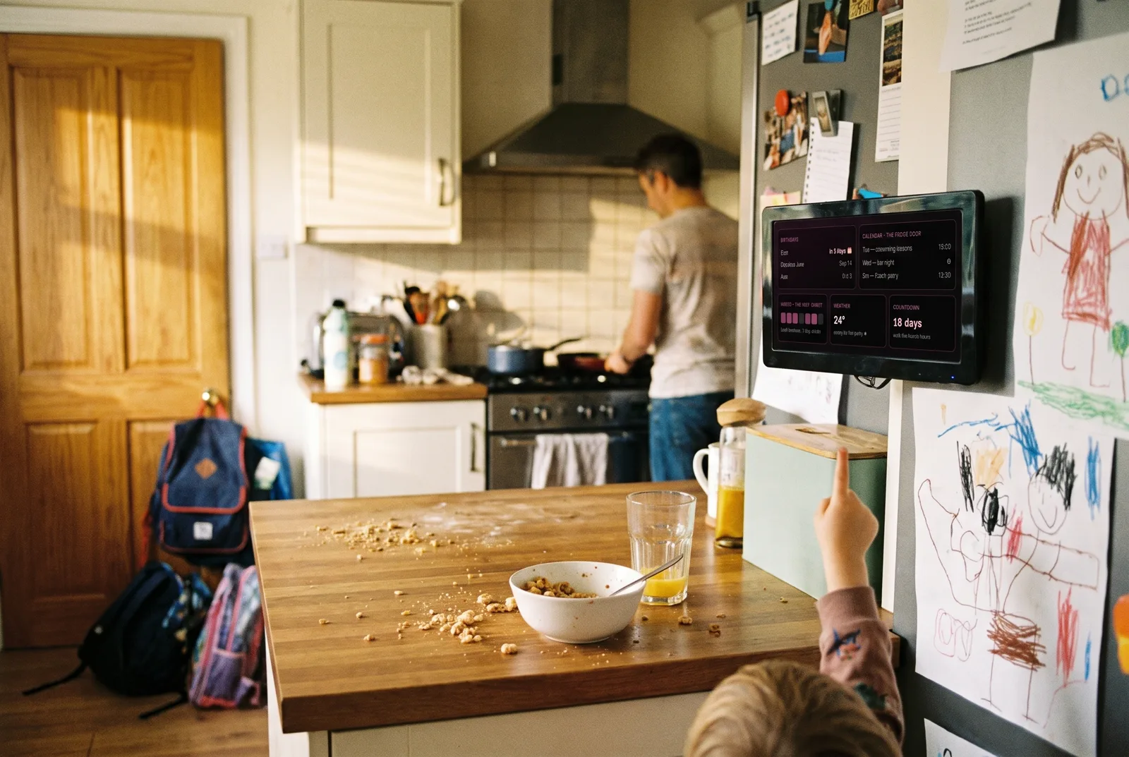 A family breakfast with a child pointing at a kitchen wall screen showing birthdays and the week ahead