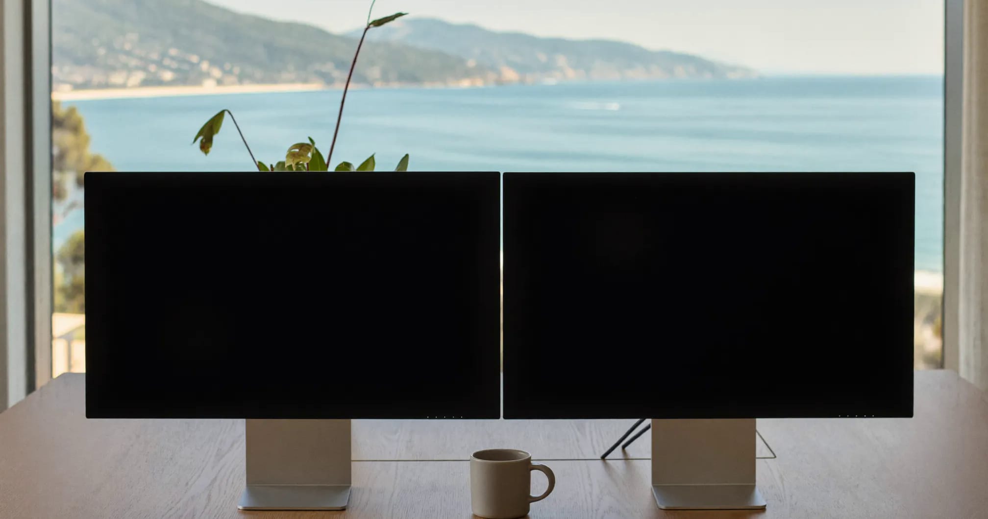 A desk in front of a window looking out over a bay and distant mountains, with two monitors on it. The left one has a browser open; the right one is running Paul's Dashboard full screen, showing the Founder screen: revenue, new customers, profit and runway across the top, then errors, deploys and uptime, then the morning brief.